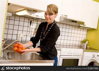A woman is washing tomatoes in the sink at home in the kitchen. The model is looking into the camera.