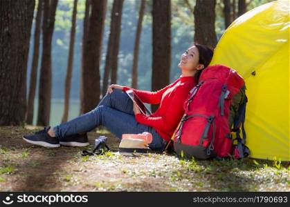 A woman leans against a yellow tent with a red backpack, She is use a tablet and enjoying nature in the midst of a beautiful pine forest beside lake, Pang Oung, Mae Hong Son, Thailand.. Women camping use tablet.