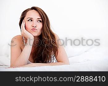 A woman lying at the end of the bed looking forward, Her hand against her chin with her other arm on the bed.