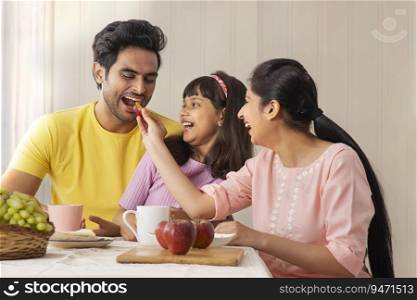 A WOMAN PLAYFULLY FEEDS HUSBAND WITH DAUGHTER HAPPILY SITTING WITH THEM