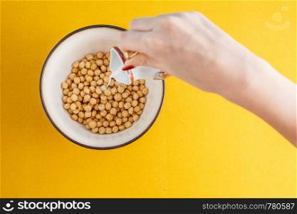 a woman's hand pours milk into honey balls from different cereals in a deep bowl
