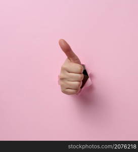 a woman&rsquo;s hand sticks out of the torn hole and shows a gesture okay, thumb up. Pink background