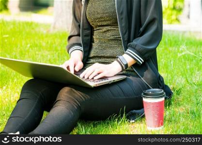 A woman sitting on the grass while using laptop computer with a red cup of coffee