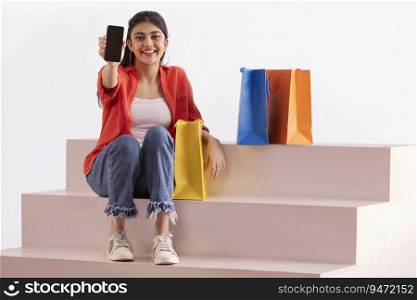 A woman sitting with colorful carrybags on stairs showing mobile phone.