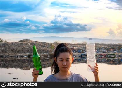 A woman stands hand holding green glass bottle and clear plastic bottle at mountain large garbage background, These waste come from urban and industrial areas can not get rid of, Pile of stink and toxic residue