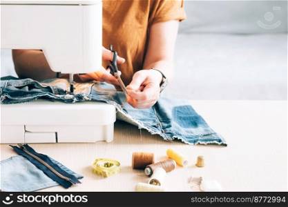 A woman tailor works at a sewing machine sews reuses fabric from old denim clothes. The concept of economical reuse of recyclable things. Homemade needlework hobby. Selective focus. A woman tailor works at sewing machine sews reuses fabric from old denim clothes