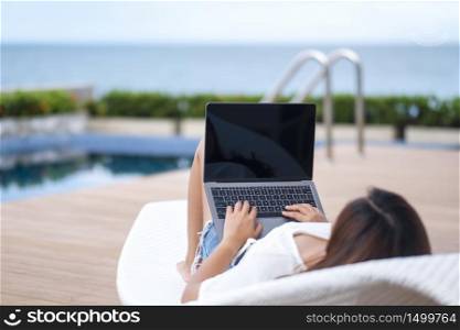A woman using and typing on laptop with blank white screen while lying down by swimming pool