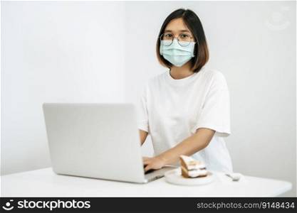 A woman wearing a mask playing on a laptop and having a cake on the plate.