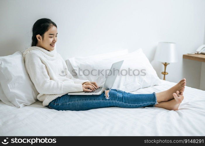 A woman wearing a white shirt on the bed and playing laptop happily.