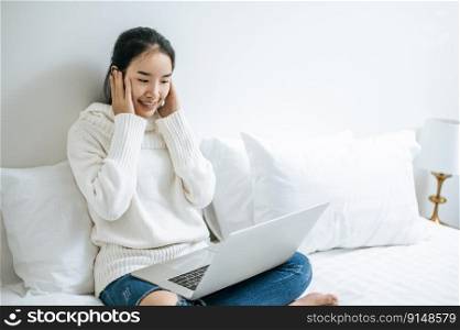 A woman wearing a white shirt on the bed and playing laptop happily.