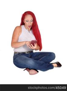 A woman with long red hair sitting in jeans on the floor shouting at hercell phone, isolated for white background.