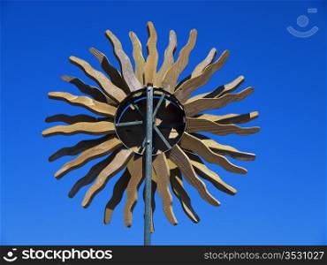 A wood whirlygig in the form of a sun with radiating rays outlined against a bright blue sky.