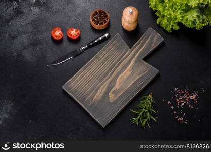 A wooden cutting board with a kitchen knife with spices and herbs on a dark concrete background. Cooking at home. A wooden cutting board with a kitchen knife with spices and herbs