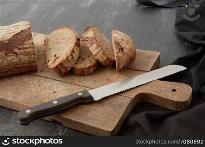 A wooden cutting board with sliced dark bread and a knife on a dark table. Fresh bread on wooden board