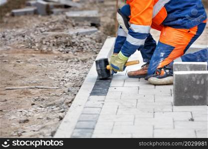 A worker in a blue and orange overalls squatting down and paving slabs on a footpath with a rubber mallet. Close-up, selective focus, copy space.. A worker lays down paving slabs with a rubber hammer in the work area.