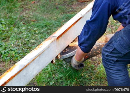 A worker in a blue overalls cuts a metal beam with an angle grinder, cutting sparks against a background of green grass. Copy space.. A worker in a blue overalls cuts a metal beam with an angle grinder.