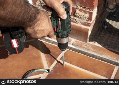 A worker is screwing a threaded rod with a drill and cordless screwdriver into a metal block previously fixed to the ground