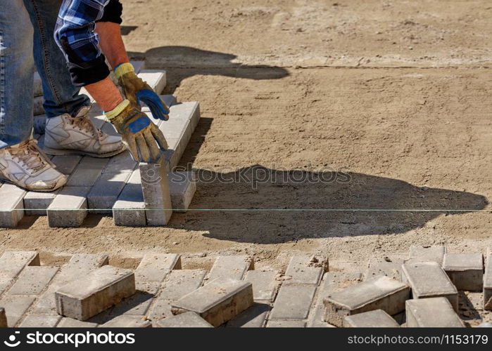 A worker lays paving slabs on a bright sunny day along a stretched cord onto prepared smooth sandy soil on the pavement, image with copy space.. A worker lays paving slabs on a prepared flat sand platform on the pavement along a stretched cord on a bright sunny day.