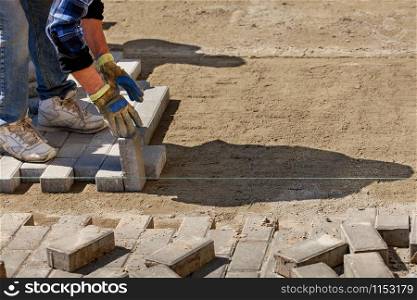 A worker lays paving slabs on a bright sunny day along a stretched cord onto prepared smooth sandy soil on the pavement, image with copy space.. A worker lays paving slabs on a prepared flat sand platform on the pavement along a stretched cord on a bright sunny day.