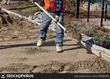A worker levels the foundation with sand and a shovel under a wooden level for laying paving slabs against the background of the workplace and preparing for work.. A worker with a shovel wears sand to level the foundation under the paving slabs.