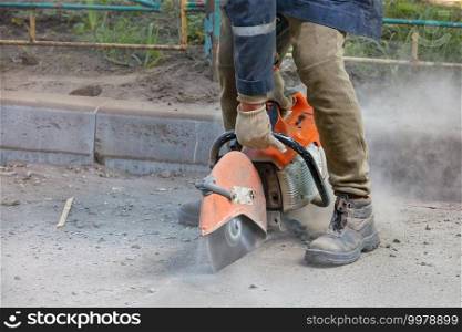 A worker uses a portable petrol saw and a diamond cutting blade to cut old asphalt in a cloud of dust, sand and flying sparks. Close-up, copy space.. A worker uses a portable chainsaw and a diamond cutting disc to cut old asphalt on the road.