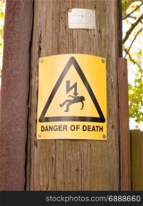 a yellow and black danger of death sign with triangle and lightening bolt on a wooden electric pole pylon outside resource