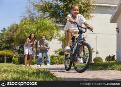 A young African American family with boy child riding his bicycle and his happy excited parents giving encouragement behind him