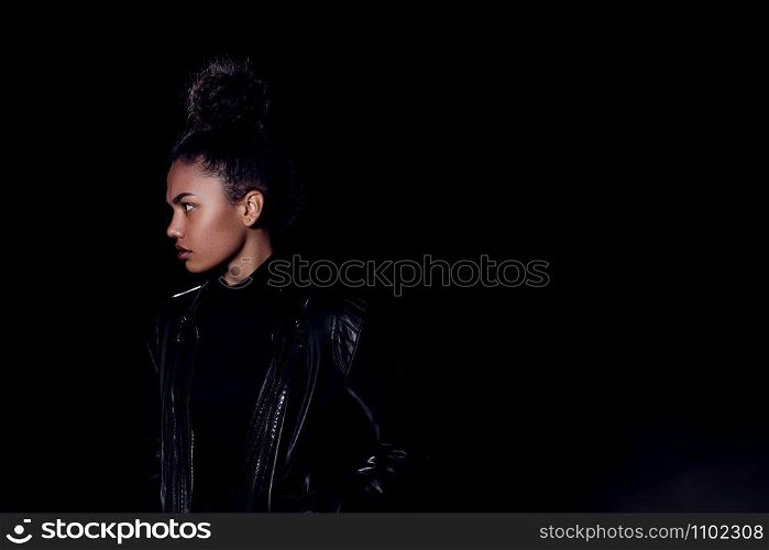 a young African American mulatto girl in a leather jacket and black clothes poses on an abandoned sand road. at night in the light of car headlights and lanterns