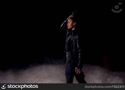a young African-American mulatto girl in a leather jacket and black clothes is walking along an abandoned sandy road with a bottle of whiskey. at night in the light of car headlights and street lights