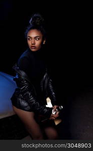 a young afro-american beautiful girl in a leather jacket,in a bodysuit and with a bottle of alcoholic beverage poses on the hood of a car. stylish street light. night.