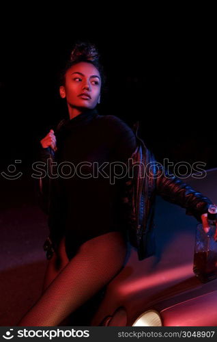 a young afro-american beautiful girl in a leather jacket,in a bodysuit and with a bottle of alcoholic beverage poses on the hood of a car. stylish street light. night.