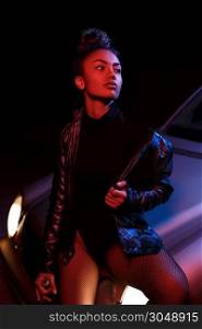 a young afro-american beautiful girl in a leather jacket,in a bodysuit and with a bottle of alcoholic beverage poses on the hood of a car. stylish street light. night.