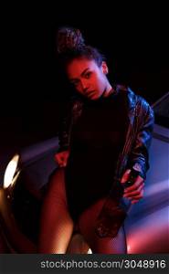 a young afro-american beautiful girl in a leather jacket,in a bodysuit and with a bottle of alcoholic beverage poses on the hood of a car. stylish street light. night.