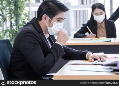 A young Asian business man is wearing mask and coughing while working in the office with blur background of colleague