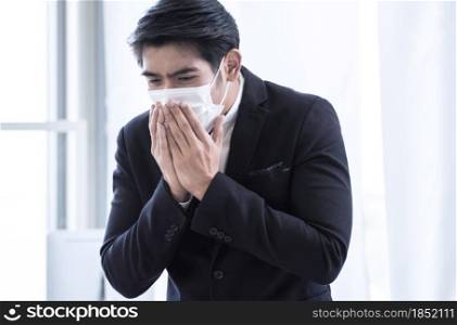 A young Asian business man is wearing mask and coughing while working in the office