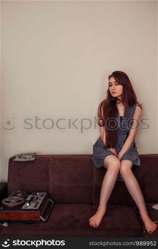 a young Asian girl in a blue striped dress sits on the couch next to reels of magnetic tape and a vintage tape recorder