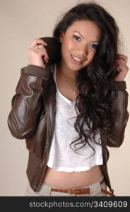 A young Asian woman in a brown leather jacked and short white t-shirtwith long brunette curly hair on beige background.