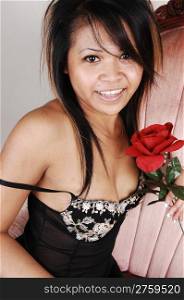 A young Asian woman in black lingerie with a red rose in her hand, sittingin a pink old armchair, smiling into the camera, over light gray background.
