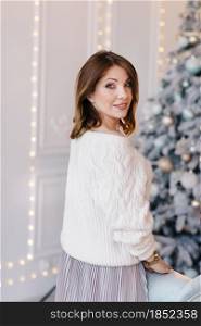 A young, attractive woman in a white sweater and skirt near the sofa near the Christmas tree and light.. A young, attractive woman in a white sweater and skirt near the sofa near the Christmas tree and light