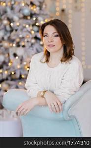 A young, attractive woman in a white sweater sits on the sofa near the Christmas tree and light.. A young, attractive woman in a white sweater sits on the sofa near the Christmas tree and light