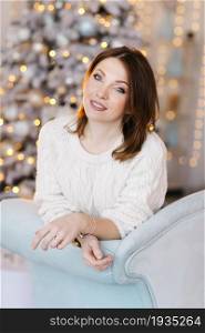 A young, attractive woman in a white sweater sits on the sofa near the Christmas tree and light.. A young, attractive woman in a white sweater sits on the sofa near the Christmas tree and light