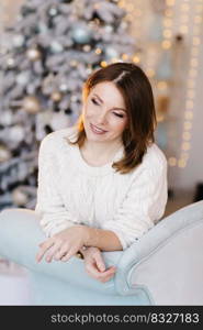 A young, attractive woman in a white sweater sits on the sofa near the Christmas tree and light.. A young, attractive woman in a white sweater sits on the sofa near the Christmas tree and light