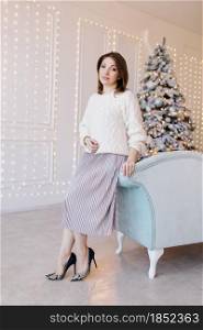 A young, attractive woman in a white sweater, skirt and high heels near the sofa near the Christmas tree and light. A young, attractive woman in a white sweater, skirt and high heels near the sofa near the Christmas tree and light.