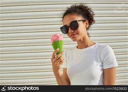 a young beautiful African-American girl in a white t-shirt and light jeans eats ice cream against a light wall on a Sunny day.