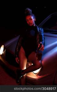 a young beautiful afro-american girl in a leather jacket, in a bodysuit posing on the hood of a car. stylish street light. night.
