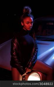 a young beautiful afro-american girl in a leather jacket, in a bodysuit posing on the hood of a car. stylish street light. night.