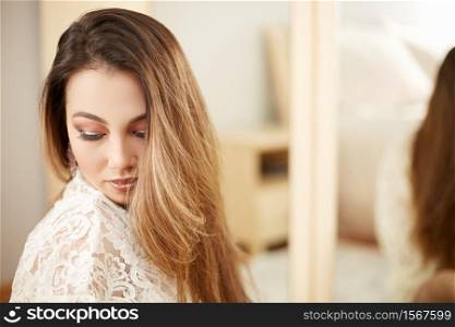 a young beautiful Asian woman in a beige lace dressing gown put on morning makeup and uses eye shadow, blush and Foundation . in the bedroom . shallow depth of focus. selective focus.