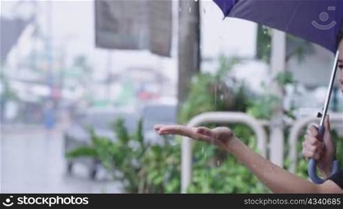 A young beautiful asian woman reaching arm to collect some waterdrops from umbrella she holding, unhappy worried female worker being late for work, hopeless sit down on street footpath, rainy season