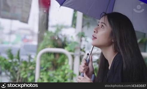 A young beautiful asian woman reaching arm to collect waterdrops from umbrella, unhappy worried worker stuck in rain, being late for appointment, hopeless sit on street footpath, heavy rainy season