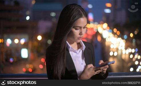 A young beautiful business woman is using smart phone in city over traffic lights at evening. business and technology concept. 